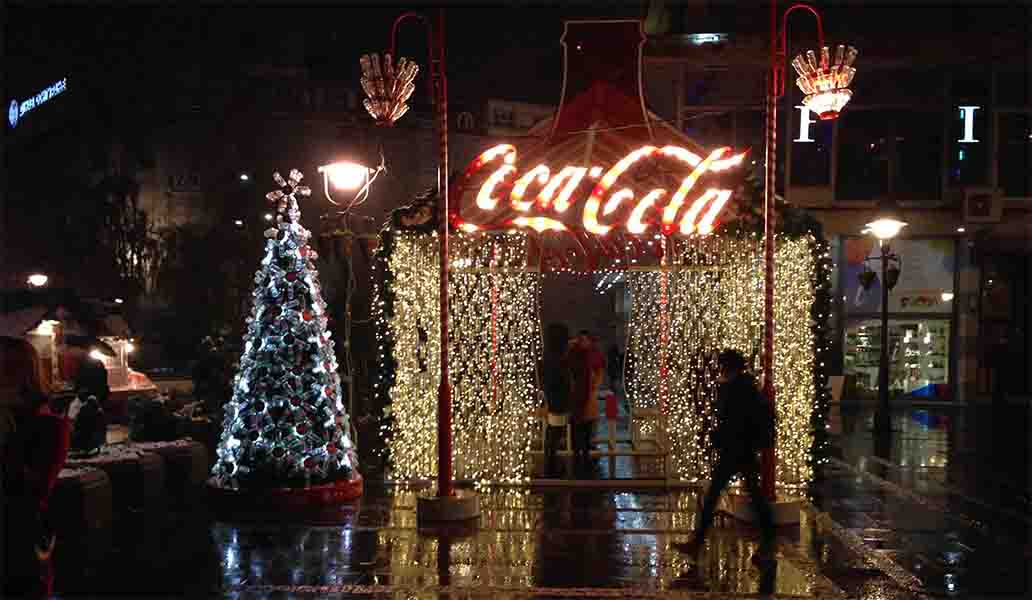 A Christmas tree, lots of lights and a Coca-Cola logo shining brightly in a dark street